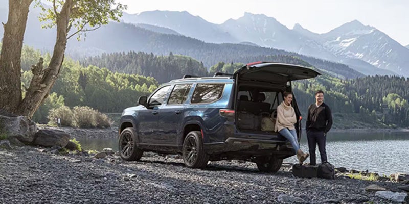 A couple by a blue SUV with its tailgate open, parked on a rocky lake shore with mountains and trees in the background.