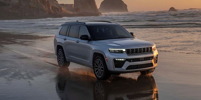 Silver Jeep Grand Cherokee L driving through shallow ocean water on a beach with cliffs in the background.