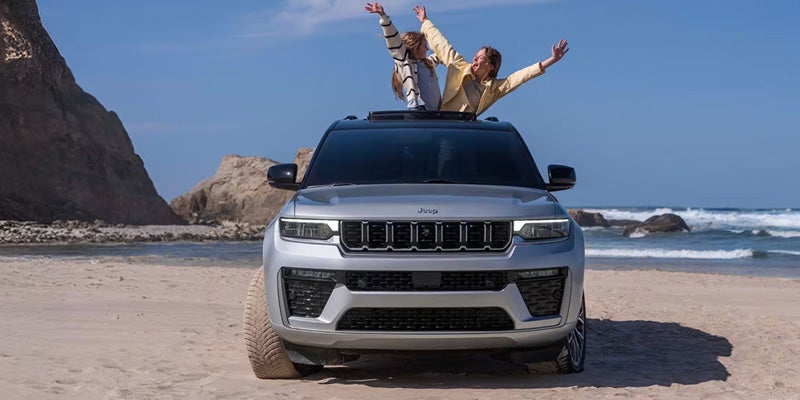 Front view of a gray Jeep with two people waving cheerfully from the sunroof on a sandy beach.