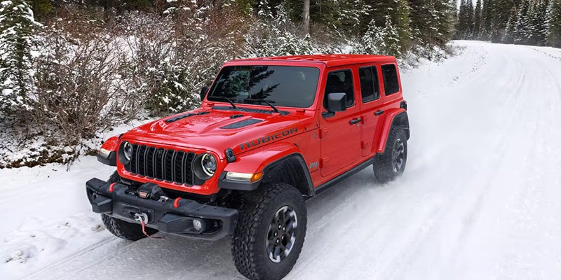 A red Jeep Wrangler Rubicon driving on a snow-covered road lined with snow-dusted evergreen trees.