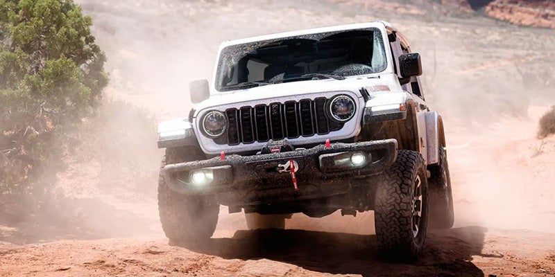 Front view of a white Jeep Gladiator Rubicon on a dusty dirt road, kicked up dust blurring the background.