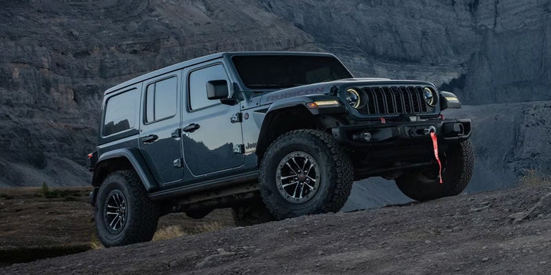 A dark blue Jeep Wrangler Rubicon with large off-road tires climbing a rocky hill with a mountain in the background.