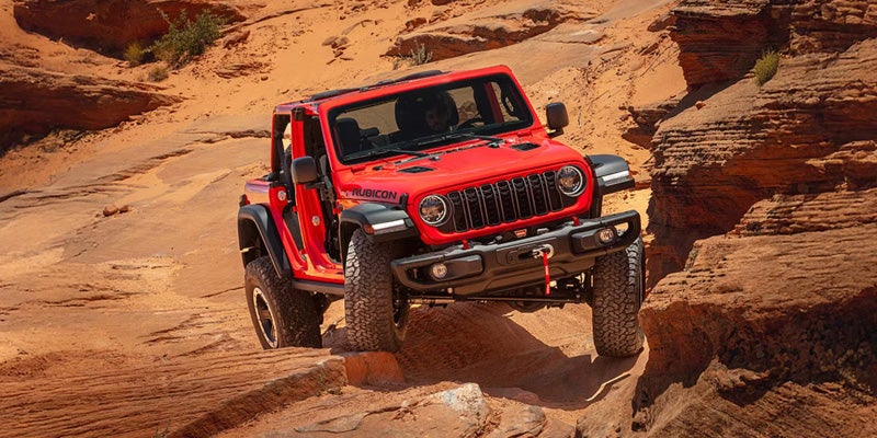 A red Jeep Rubicon Wrangler off-roading on a rocky, sandy trail with sandstone formations.