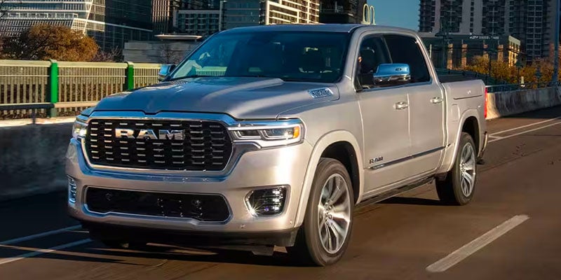 A silver Ram pickup truck with a large grille and "RAM" lettering drives down a road, with city buildings in the background.