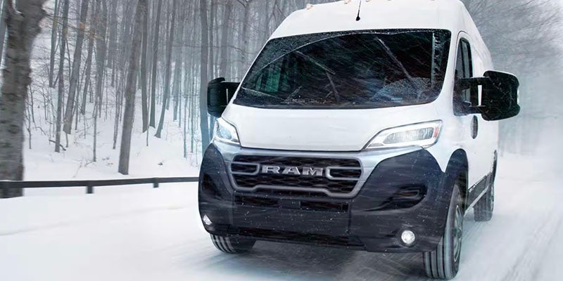 White Ram ProMaster van driving on a snow-covered road through a bare winter forest.