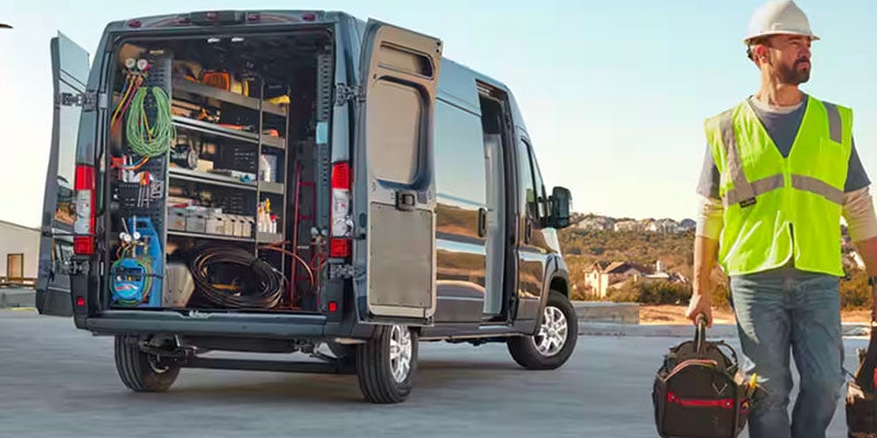 A technician in a hard hat and safety vest walks away from an open cargo van filled with tools and equipment.