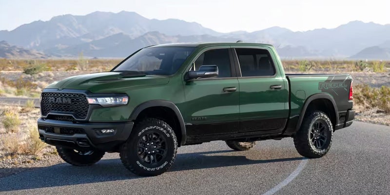 A dark green Ram Rebel truck with black wheels and tires, parked on a paved road with mountains in the background.
