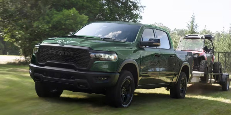 A green Ram pickup truck drives left on a grassy field, towing a red and black side-by-side on a trailer.