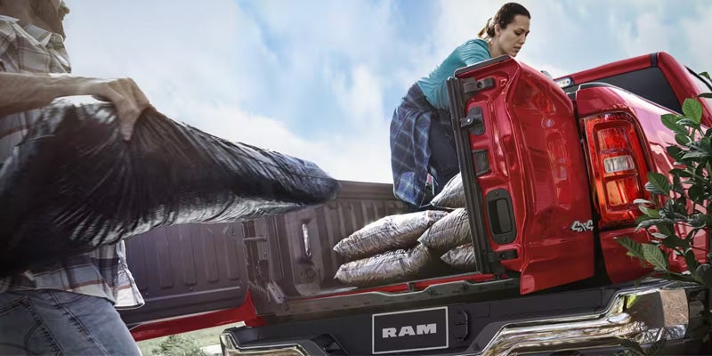 A man loads a black bag into the bed of a red Ram pickup truck, while a woman arranges other bags in the bed.