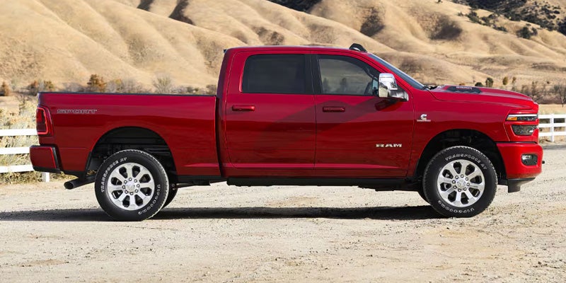A shiny red Ram pickup truck with chrome wheels is parked on a dirt path with dry hilly terrain in the background.