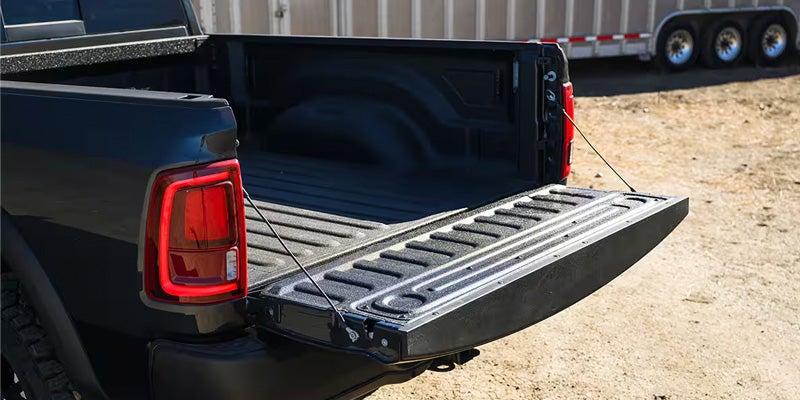 The tailgate of a dark gray pickup truck is open, revealing a black bedliner, with a trailer in the background.