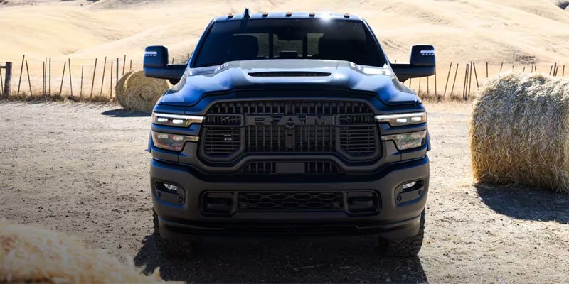 Front view of a dark blue Ram truck with black grille and headlights, parked on a dirt road next to hay bales.