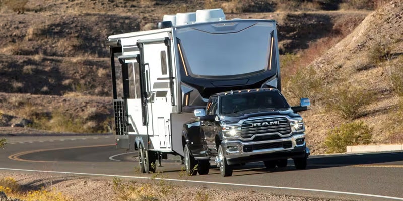 A dark grey Ram truck with a silver front grille pulling a large fifth-wheel RV on a winding desert road.