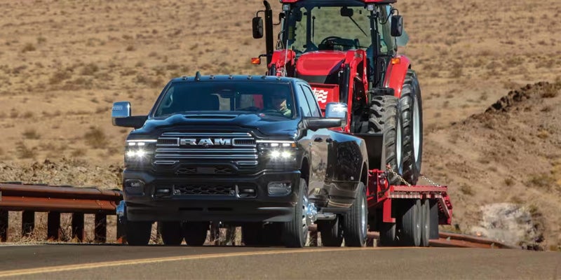 Dark gray pickup truck towing a red tractor on a flatbed trailer on a desert highway.