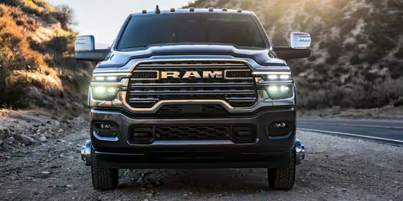 Front view of a dark grey Ram truck with its headlights on, parked on a dirt road next to a highway at dusk.