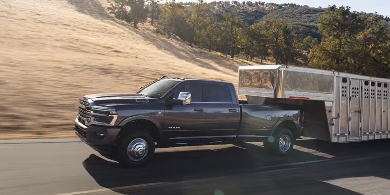 A dark gray Ram truck with a silver livestock trailer on a highway, with dry hills and trees in the background.
