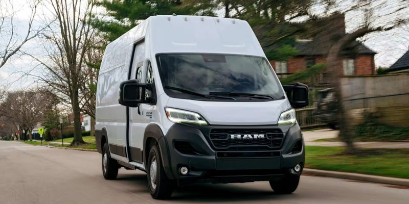 A white Ram ProMaster van with a black grille and bumper drives down a street with houses and trees.