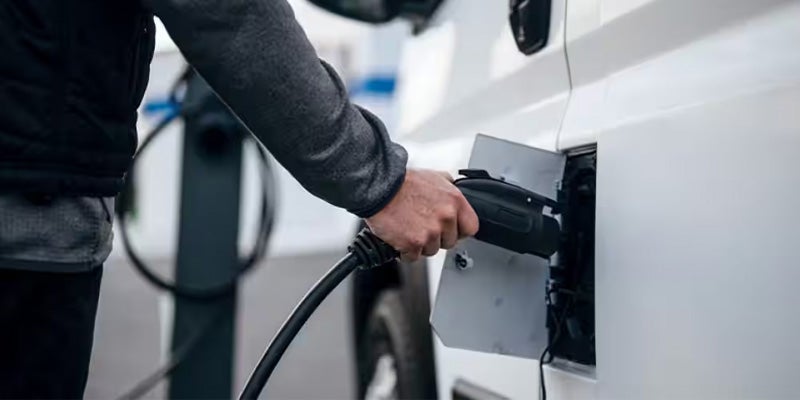 A person's hand plugging an electric vehicle charging cable into a white van at a charging station.
