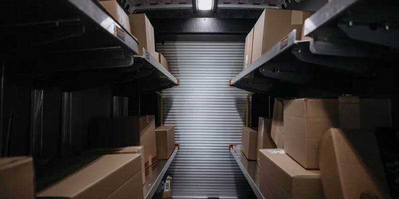Interior of a delivery truck or van, with shelves full of cardboard boxes and a closed roll-up door at the back.
