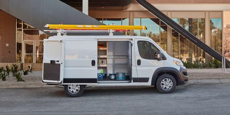White work van with open side and back doors, exposing internal shelving and tools, parked on asphalt.