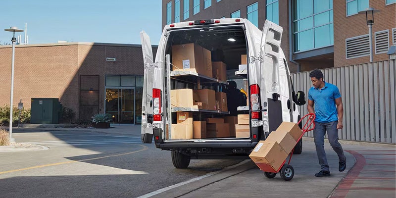 A man unloads boxes from a white cargo van with its rear doors open, putting them onto a hand truck.