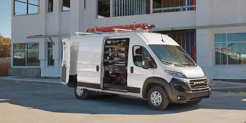 White Ram ProMaster cargo van with open side door showing tool shelves and a ladder on the roof, parked outside a building.