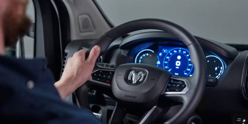 A driver's hands on a modern Ram steering wheel with controls and a digital dashboard with blue screen.