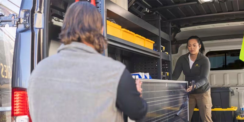 Two people, one man, one woman, load a solar panel into the back of a large white work van fitted with shelves.