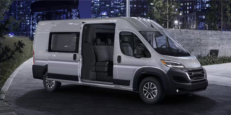 Silver Ram ProMaster passenger van, with open sliding door and rear doors, parked on a city street at night.